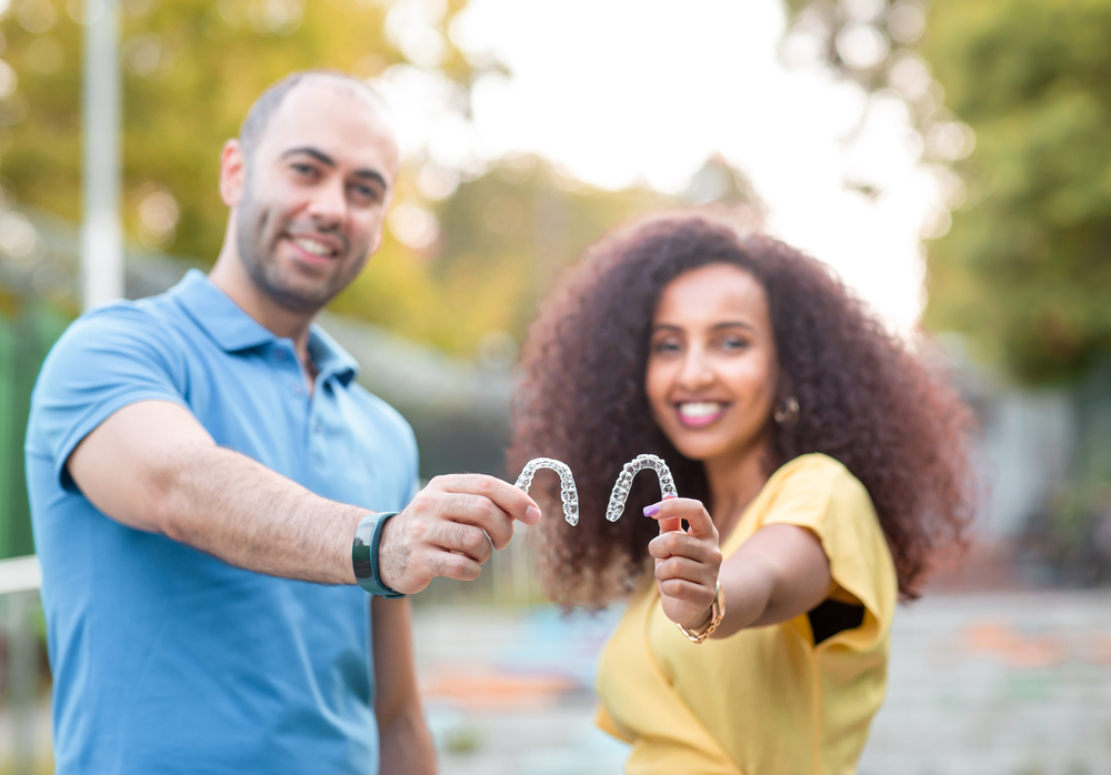 Happy Couple Showing Invisalign Aligners – Invisalign in Irvine Smiling couple outdoors holding clear Invisalign aligners together, showcasing their straight smiles – Invisalign in Irvine