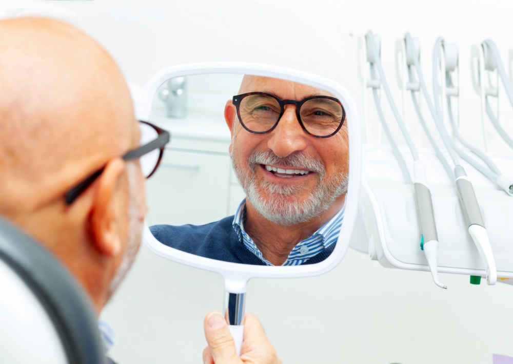 Happy Patient Admiring New Smile – Dental Implants Irvine Smiling man looking in the mirror after receiving dental implants – Dental Implants Irvine