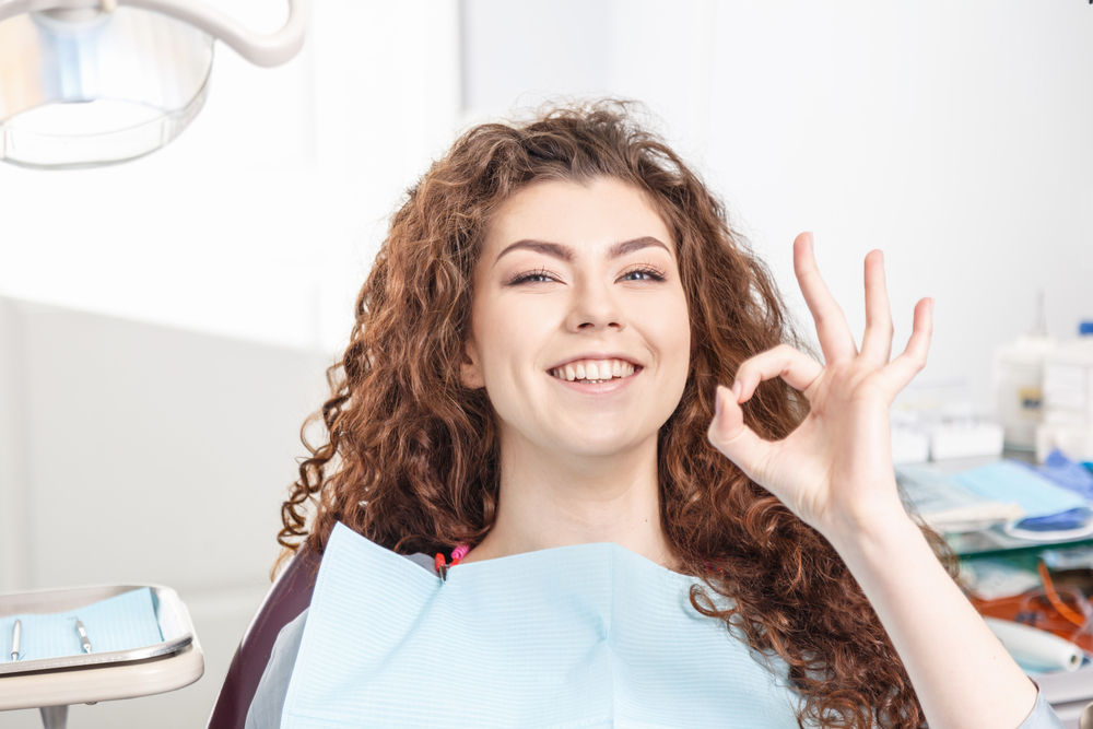Happy Patient After Dental Implant Procedure Smiling woman sitting in a dental chair showing an OK sign after receiving dental implants at the dentist’s office - Dental Implant