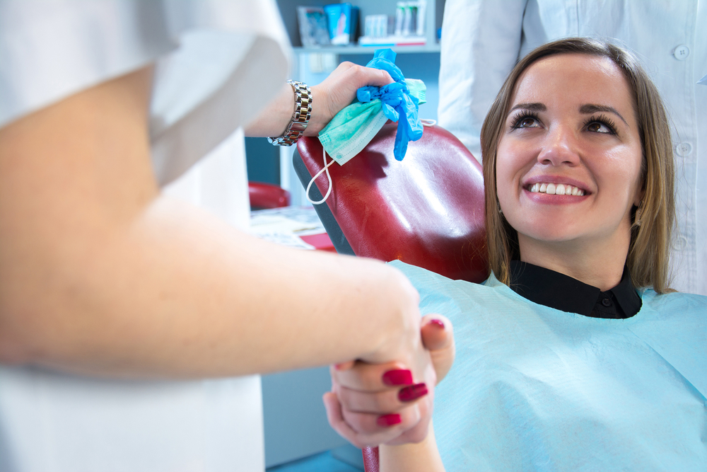 A smiling woman sits in a red dental chair wearing a blue bib, holding hands with a dental professional, conveying comfort and trust in a clinical setting – Anderson Family Dental