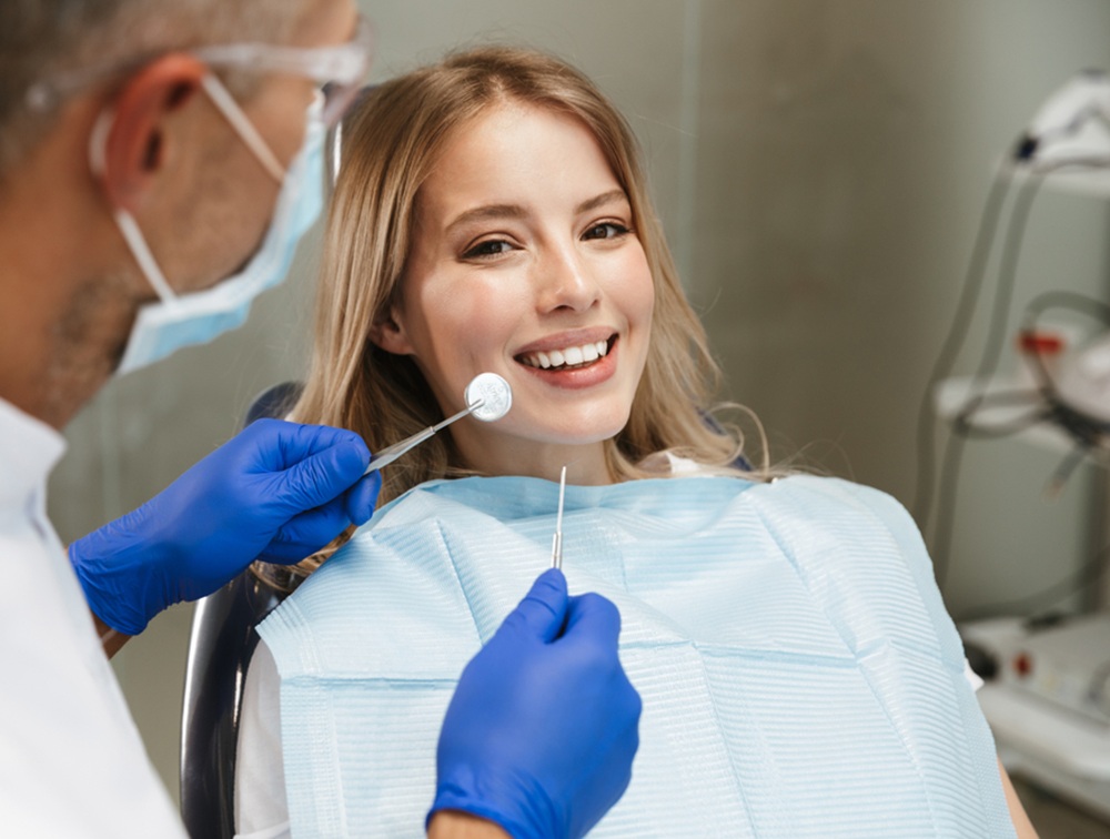 Routine Dental Checkup – Lake Forest Dentist Female patient smiling during a dental exam while the dentist checks her teeth with dental tools – Lake Forest Dentist