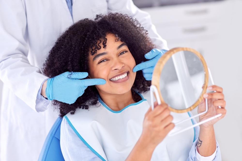 Smiling Woman Checking Her Whiter Smile at Dentist – Teeth Whitening in Irvine Woman sitting in a dental chair holding a mirror and smiling as a dentist points to her bright teeth after whitening treatment – Teeth Whitening in Irvine