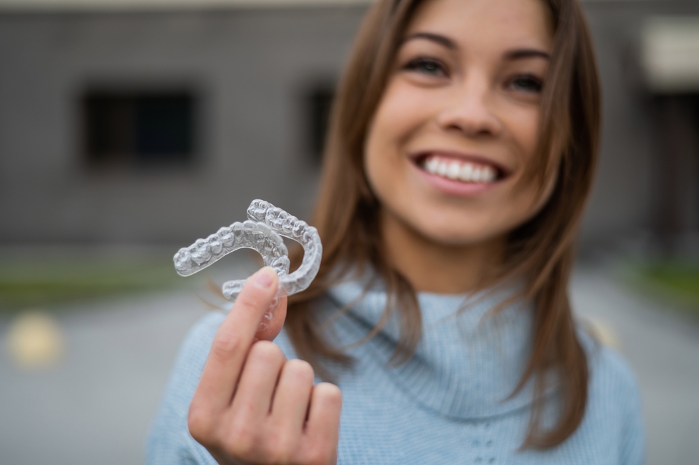 Woman Holding Clear Aligners and Smiling – Invisalign in Irvine Close-up of a smiling woman showing transparent Invisalign aligners outdoors – Invisalign in Irvine